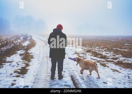 Mann mit Labrador Retriever Hund beim Wandern auf dem Land im Winter. Felder bedeckt mit dem ersten Schnee Stockfoto