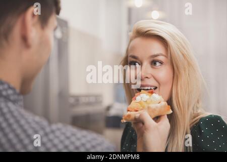 Schöne junge Frau genießen Beißen leckere Pizza, ihr liebevoller Freund offerng sie. Mann füttert Pizza zu seiner schönen Frau. Glücklich liebende Paar Stockfoto