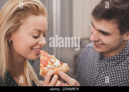 Nahaufnahme eines glücklichen Paares teilen Pizza, mit romantischen Abendessen zu Hause. Wunderschöne Frau lächelt, ihr Freund bietet ihr Stück Pizza. Gut Aussehend Stockfoto