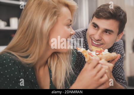 Nahaufnahme einer glücklichen schönen Frau, die ihren hübschen Mann anlächelt und zu Hause leckere Pizza isst. Schöner Mann beobachten seine schöne Freundin essen ta Stockfoto