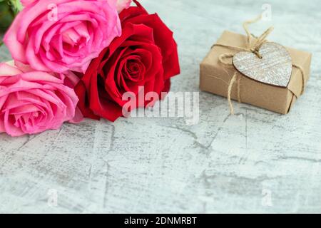 Geschenk in Bastelpapier neben Strauß Rosen auf weißem Stein Hintergrund gewickelt. Romantisches Geschenk zum valentinstag mit Platz für Text. Konzept des Heiratsvorschlags. Weichfokus. Speicherplatz kopieren Stockfoto