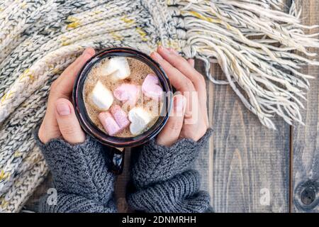 Weibliche Hände halten Tasse mit einem heißen Getränk mit Marshmallows auf der Holztisch mit warmen Strick Wollschal. Weihnachtshintergrund - das Konzept von Winter, Wärme, Urlaub und Veranstaltungen.Weiche Fokus.Draufsicht Stockfoto