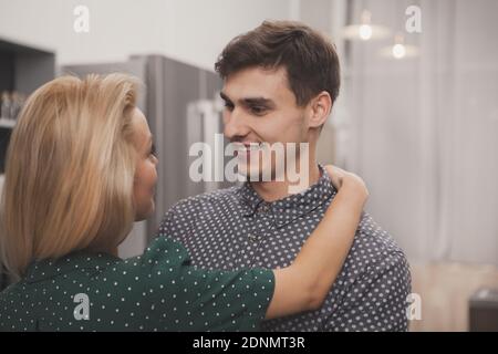 Schöner junger Mann lächelt freudig seine schöne Frau an und umarmt sie in der Küche zu Hause. Glücklicher Mann umarmt seine liebevolle Frau, kopieren Raum. Relat Stockfoto
