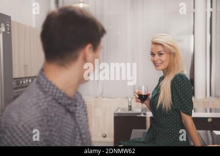 Wunderschöne Frau sitzt auf dem Tisch in der Küche, Wein, Blick auf ihren Mann verführerisch. Liebevolles Paar genießen romantische Nacht zu Hause. Sensua Stockfoto