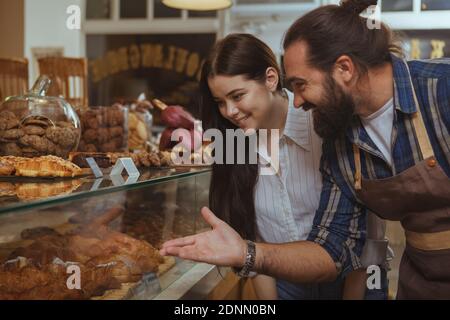 Junge schöne Frau kauft köstliche frisch gebackene Gebäck in der lokalen Bäckerei. Professioneller Bäcker zeigt auf das Display und spricht mit seinem Kunden. Bär Stockfoto