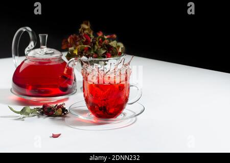 Herbstabend Teezeit mit Spritzer in roter Kräuterteetasse, transparente Teekanne mit Weißdornbeeren, Hagebutte, trockene Blätter auf weißem Holztisch, schwarz w Stockfoto