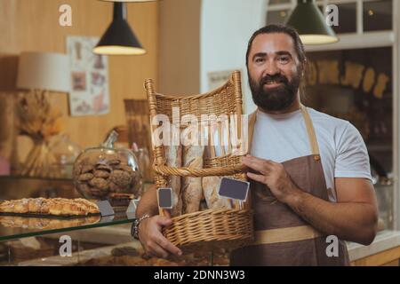 Glücklich gut aussehende bärtige Bäcker trägt Schürze, Korb mit frischem Brot, Kopie Raum. Reif männlich Bäcker genießen arbeiten bei seinem Café, se Stockfoto