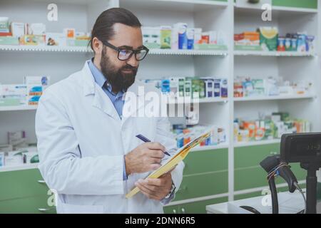 Reifer bärtiger Apotheker, der Papiere füllt und in seiner Drogerie arbeitet. Erfahrener Chemiker in Laborkittel, der hinter dem Kassenschalter in der Apotheke steht. Stockfoto