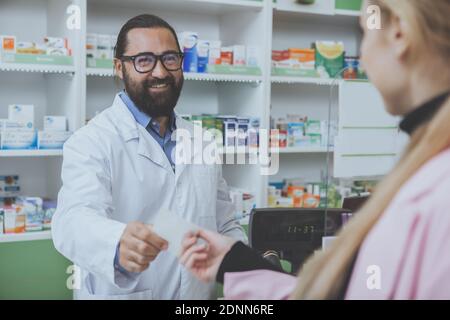 Fröhlicher bärtiger Apotheker, der in der Drogerie arbeitet und seinem weiblichen Kunden verschreibungspflichtiges Papier gibt. Unkenntlich Frau Kauf Medikamente bei der Stockfoto