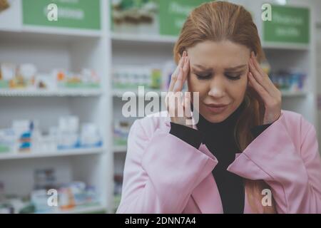 Nahaufnahme einer jungen attraktiven Frau mit schrecklichen Kopfschmerzen, Apotheke im Hintergrund. Frau mit Migräne reiben Schläfen, Shopping für Painkille Stockfoto