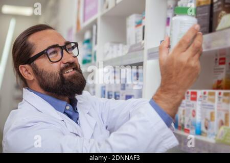 Schöner bärtiger reifer Apotheker, der in seiner Drogerie arbeitet und medizinische Produkte in den Regalen organisiert. Fröhlicher männlicher Chemiker prüft Vorrat in einem ai Stockfoto