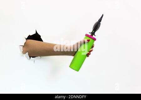 Eine weibliche Hand mit einer roten Maniküre durch zerrissenes weißes Papier hält eine Ballonpumpe mit grünem Griff. Hand durch zerrissenes weißes Papier Stockfoto