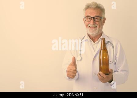 Studio shot von Happy älterer bärtiger Mann Arzt lächelnd, während sie eine Flasche Bier und den Daumen auf weißem Hintergrund Stockfoto