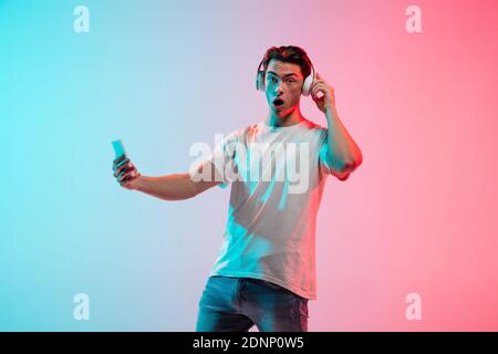 Musik hören, schockiert. Das Porträt des jungen Kaukasiermannes auf einem blau-rosa Hintergrund in Neon. Konzept der Jugend, menschliche Emotionen, Gesichtsausdruck, Verkauf, Anzeige. Halbe Länge, Copyspace. Stockfoto