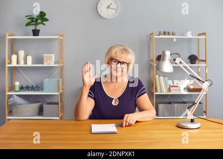 Positive ältere Frau grüßt jemanden mit der Hand und schaut an Kamera während eines Online-Meetings Stockfoto