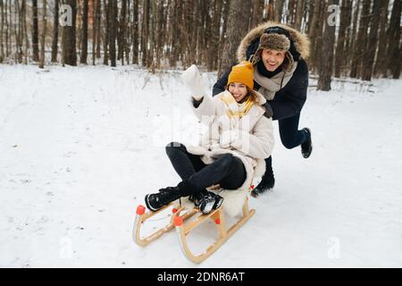 Mann schiebt mit seiner Freundin einen Holzschlitten in einen Wald und nimmt Geschwindigkeit auf Stockfoto