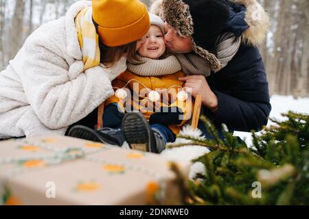 Glückliche Eltern küssen ihr kleines Mädchen, sitzen auf einem Schlitten. In einem Winterwald Stockfoto