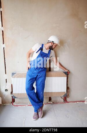 Volle Länge der bärtigen jungen Mann Klempner Überprüfung Heizkörper während der Installation Heizsystem in der Wohnung. Männlicher Arbeiter trägt blaue Arbeitskleidung und Schutzhelm, steht in der Nähe der Heizbatterie. Stockfoto