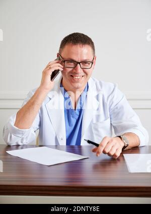 Glücklicher Mann Chirurg in einer Brille lächelnd, während er am Tisch mit Papieren sitzt. Männlicher Arzt hält Stift und spricht auf Handy in der Klinik. Konzept der Medizin, Kommunikation und Gesundheitswesen Arbeitnehmer. Stockfoto