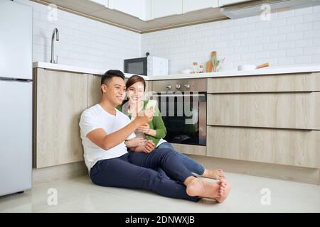 Liebe liegt in der Luft. Schönes junges Paar, das Kaffee und Orangensaft trinkt, während es zu Hause auf dem Küchenboden sitzt