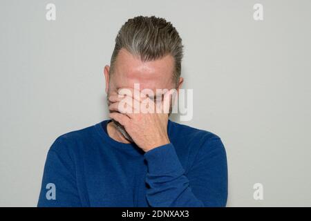 Despondent middle-aged man holding his head in his hand covering his eyes as he looks down dejectedly against a white studio wall Stock Photo