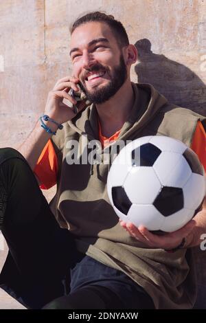 Gutaussehender Sportler lacht und telefoniert, während er sich mit seinem Fußballball, Technikkonzept und urbanem Sport-Lifestyle in der Stadt ausruht Stockfoto