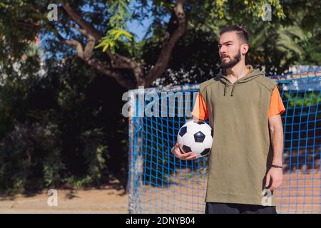 Schöner Hipster-Fußballspieler vor dem Tor eines städtischen Platzes mit dem Ball unter dem Arm, Konzept des gesunden Lebensstils und des urbanen Sports in t Stockfoto