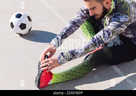 Der junge Sportler streckt die Beine auf dem Boden neben seinem Fußballball in einem Betonfußballplatz, Konzept des gesunden Lebensstils und Stadtspor Stockfoto