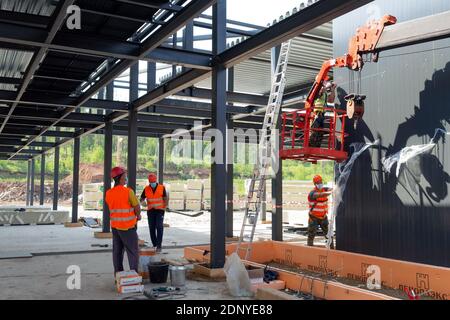 Ein Installateur in einer Hängewiege und ein Arbeitsteam installieren Wandpaneele auf der Baustelle eines medizinischen Hilfs- und Rettungszentrums. Stockfoto