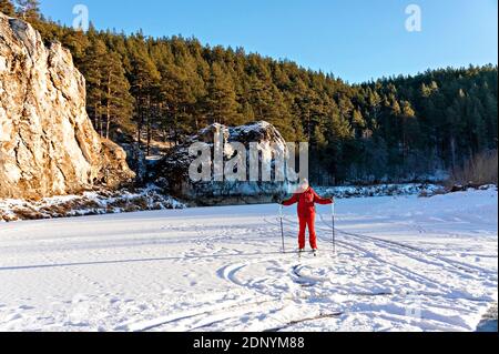 Junger Mann in roten Winterkleidung Skifahren entlang der gefrorenen verschneiten Fluss in der Nähe von schönen Klippen mit Pinienwald. . Winteraktivitäten und Spaß, Sport, aktiv Stockfoto