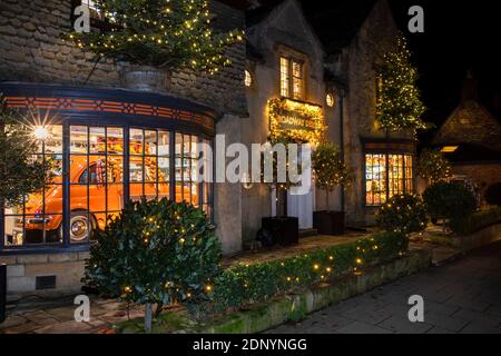 Großbritannien, Gloucestershire, Broadway, High Street, Broadway Deli beleuchtet für Weihnachten, mit Auto im Schaufenster Stockfoto