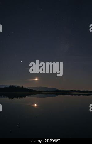 Wachender Halbmond hinter Lichtwolken, Sternen und Milchstraße, die sich in der Abenddämmerung im Kirchsee spiegeln, Bayern, Deutschland Stockfoto