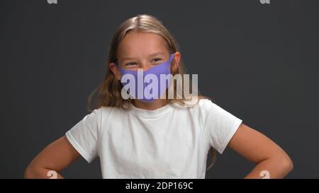 Schulmädchen trägt blau wiederverwendbare Gesichtsmaske trägt weißes T-Shirt lächelnd mit ihren Augen Blick auf die Kamera, während im Studio stehen. Isoliert auf Schwarz Stockfoto