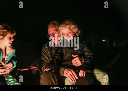 Enkelin sitzt auf dem Schoß des Großvaters und sitzt um die Feuerstelle herum Stockfoto