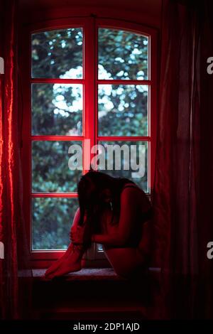 Frau sitzt mit umarmenden Knien an der Fensterbank Stockfoto