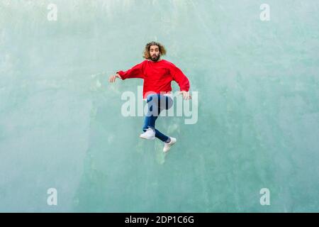 Porträt des jungen Mannes mit rotem Sweatshirt, das vor der grünen Wand in die Luft springt Stockfoto