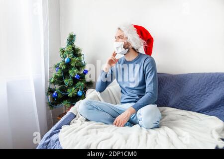 Covid-19 Weihnachten und Pandemie Neues normales Konzept. Junger Mann mit Gesichtsmaske und Weihnachtsmütze, der auf der Couch im weihnachtlich dekorierten Zimmer sitzt. Stockfoto
