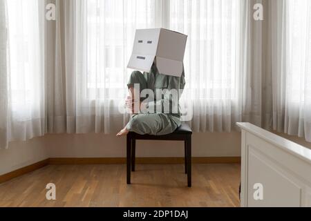 Frau trägt einen Karton auf dem Kopf mit gelangweilten Smiley sitzen Auf dem Stuhl Stockfoto
