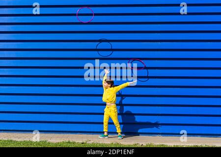 Kleines Mädchen trägt gelbe Trainingsanzug Werbungen Gymnastik Ringe in der Luft vor blauem Hintergrund Stockfoto