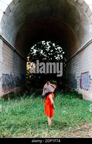 Kleines Mädchen mit einem Pferdekopf und einem roten Kleid Stehen in der Natur unter einer Brücke Stockfoto