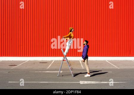 Junge Männer und Frauen, die mit einer Leiter vor sich auftreten Einer roten Wand Stockfoto