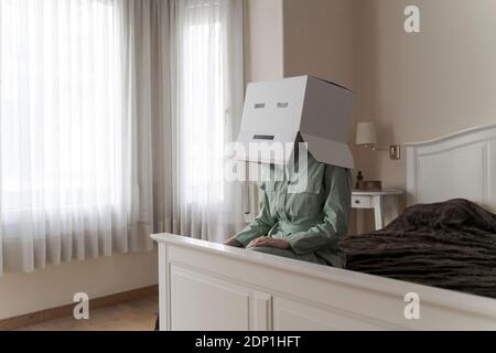 Frau trägt einen Karton auf dem Kopf mit gelangweilten Smiley sitzen Auf dem Bett Stockfoto