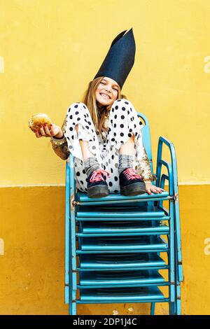 Portrait von lachenden Mädchen mit schwarzen Krone sitzen auf Stapel Stühle essen Hamburger Stockfoto