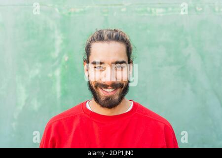 Porträt eines bärtigen jungen Mannes, der vor der grünen Wand rotes Sweatshirt trägt Stockfoto