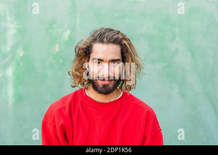 Porträt eines bärtigen jungen Mannes, der vor der grünen Wand rotes Sweatshirt trägt Stockfoto
