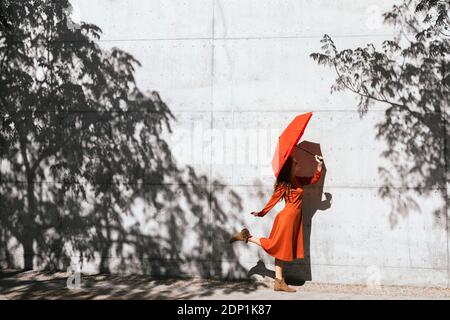 Frau in rotem Kleid hält roten Regenschirm, während Sie gegen stehen Schattenmauer von Bäumen Stockfoto