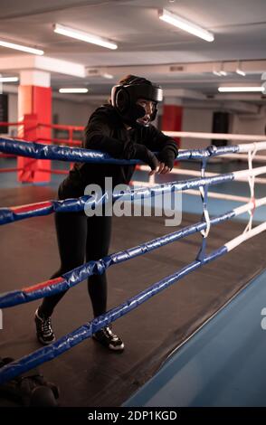 Ganzkörper müde Sportlerin in Schutzhelm und Sportswear lehnend An Seilen und nach dem Kampf auf Boxring ruhen Stockfoto