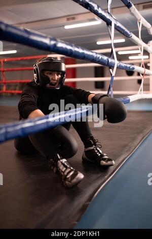 Full body aggressive female boxer in protective gear screaming while sitting on boxing ring and resting after fight Stock Photo