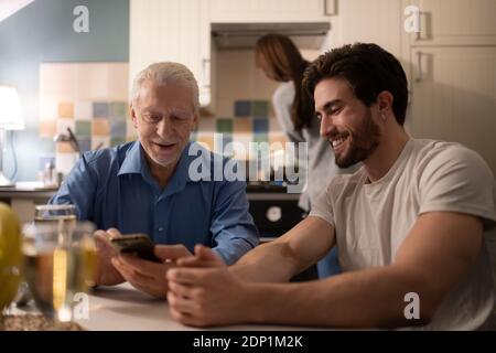 Senior father using smartphone and showing photos to cheerful young son while sitting at kitchen table near cooking woman before family dinner Stockfoto
