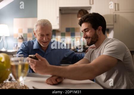 Fröhlicher älterer Mann unterstützt bärtigen Sohn zeigt Fotos auf dem Smartphone Während der gemeinsamen Zeit beim Familienessen Stockfoto